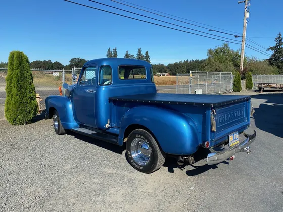 Blue 1950s Chevrolet pickup with a tonneau cover and chrome bumper parked on gravel in the sun