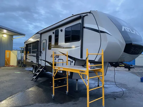 Tan Atlas fifth wheel with a yellow rolling scaffold set up beside it on wet pavement at dusk