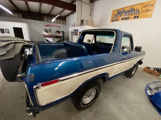 Blue and white late 1970s Ford Bronco with the top off and a tire mounted on the back, inside the shop