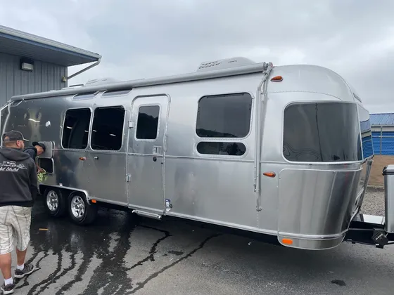 Two men wiping down the polished aluminum side of an Airstream trailer on wet pavement
