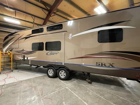 Tan Cougar SRX fifth wheel in the shop with blue tape along the lower edge and a scaffold at the rear