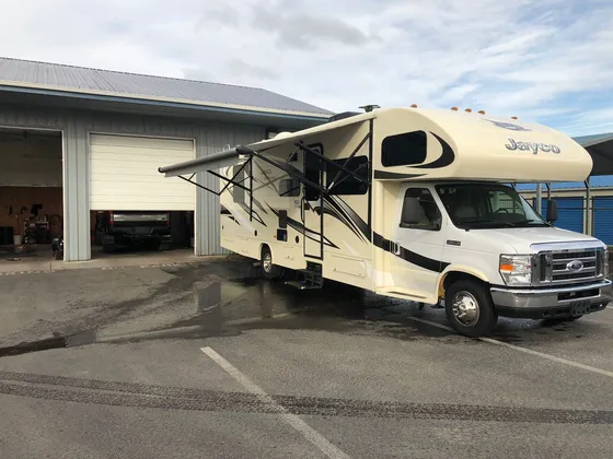 Cream Jayco Class C motorhome with its awning out, parked on wet pavement outside the shop