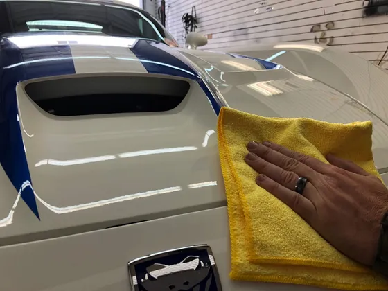 Hand with a yellow microfiber towel on the hood of a white sports car with blue stripes and a snake badge