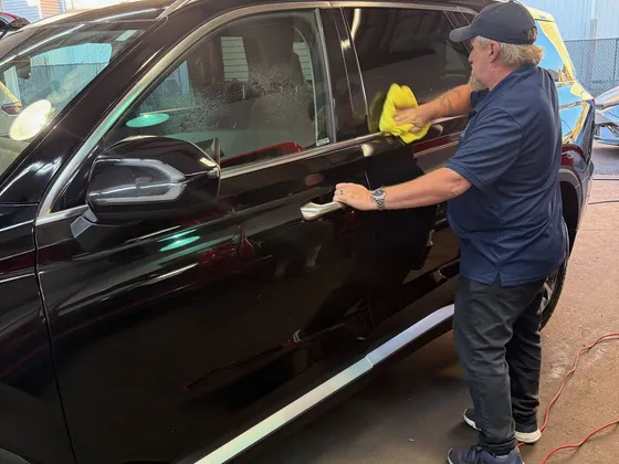 Worker in a navy polo wiping the driver side door of a dark SUV with a yellow towel in the shop