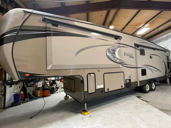 Tan Eagle Premier fifth wheel with dark swoosh graphics parked inside the shop, curb side