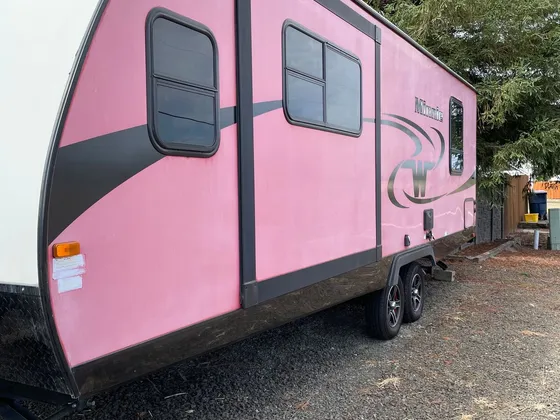 Faded pink travel trailer with dark graphics and a Minnie badge, parked on gravel