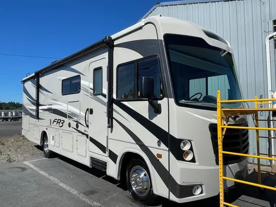 Tan and black FR3 Class A motorhome parked next to the shop wall with yellow scaffolding in front