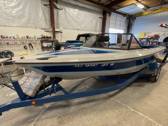 Older gray and blue ski boat with a windshield frame on a blue trailer inside the shop