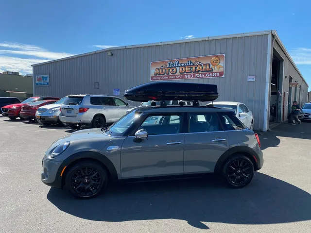 Gray four-door Mini Cooper with black wheels and a roof cargo box parked in front of the shop
