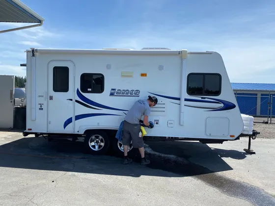 Worker bent over the side wall of a white Lance travel trailer with a buffing pad and spray bottle