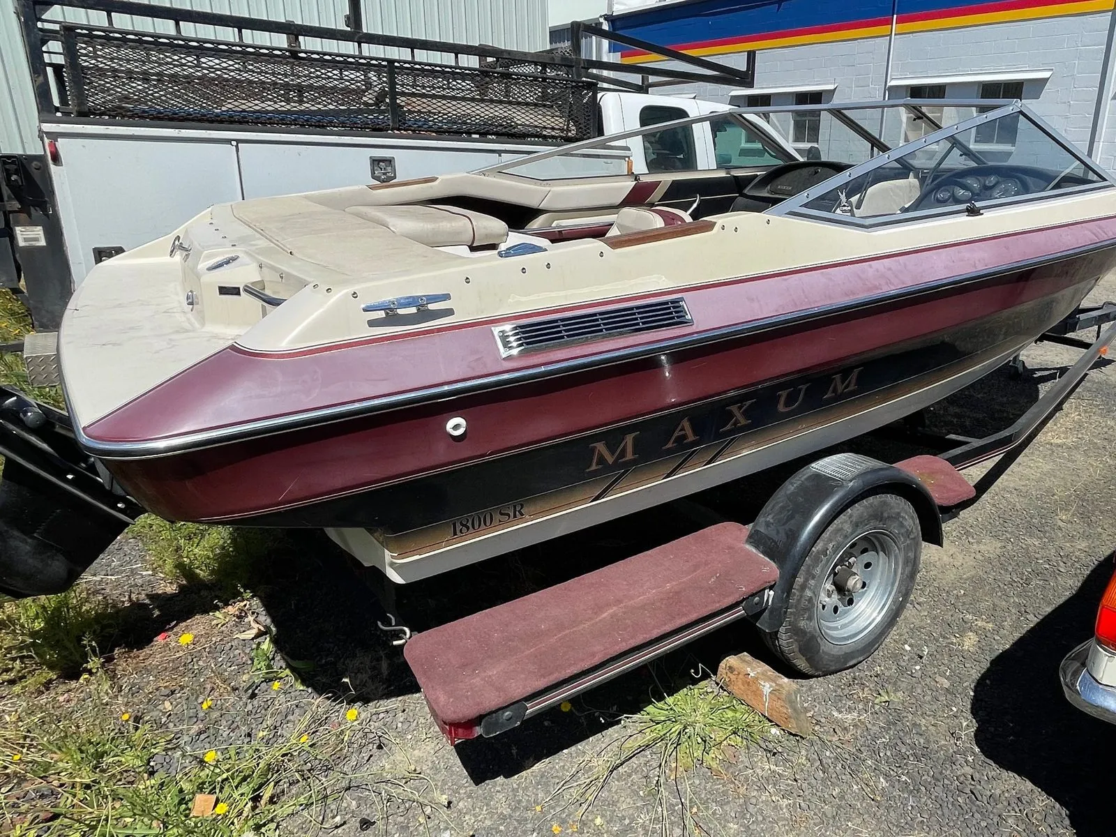 Maroon and tan Maxum 1800 SR runabout with a chalky dull deck sitting on a trailer in a gravel lot