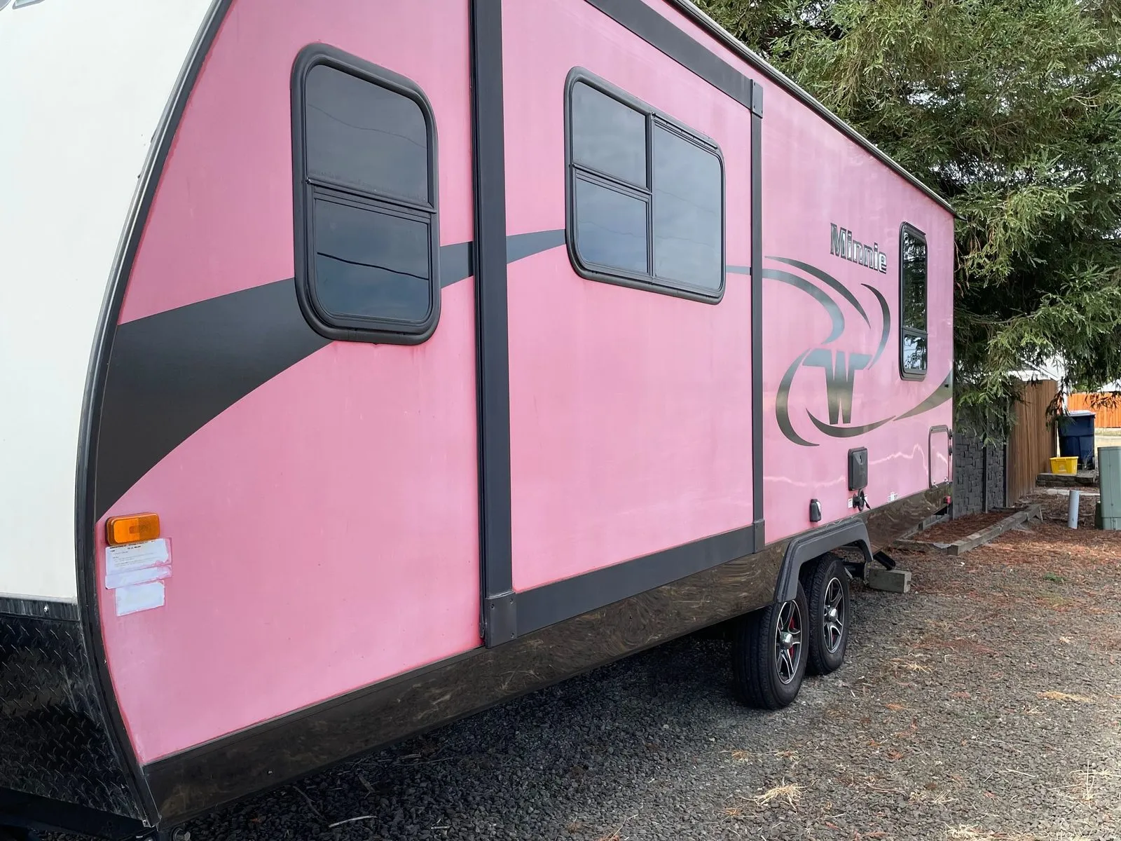 Faded pink travel trailer with dark graphics and a Minnie badge, parked on gravel