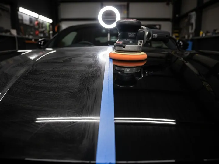 Polisher on a black hood with swirl marks on one side of the tape line and polished paint on the other