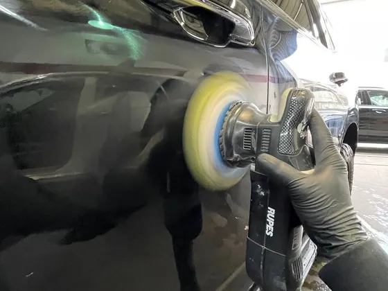 Gloved hand holding a Rupes polisher with a yellow foam pad against a black SUV door