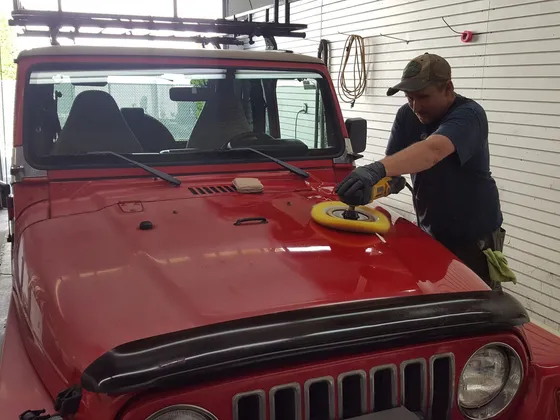 Worker running a yellow polishing pad over the hood of a red Jeep Wrangler in a shop bay