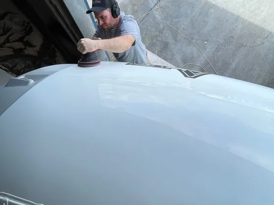 Man in a ball cap and headphones running a dual-action polisher on the roof cap of a gray RV
