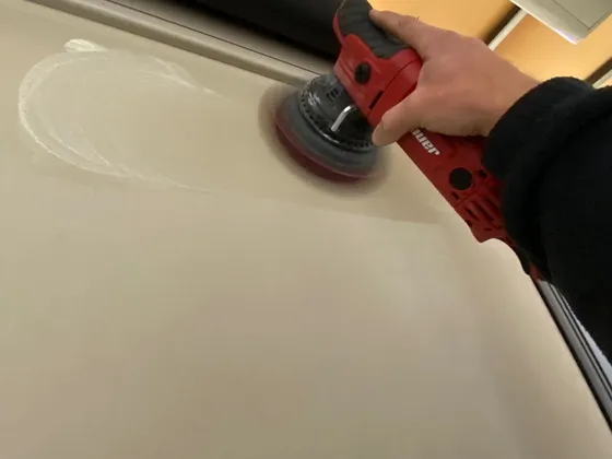 Hand holding a red dual action polisher on a tan RV wall with a swirl of compound on the surface