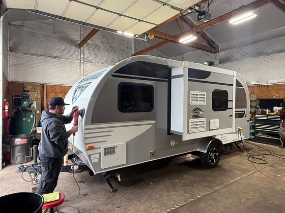 Man polishing the front of a small gray and white travel trailer with a red polisher inside the shop