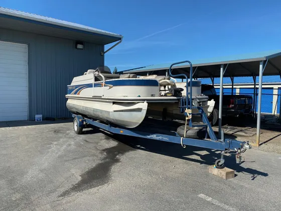 Cream and blue pontoon boat with tan seats sitting on a trailer outside the shop on a sunny day