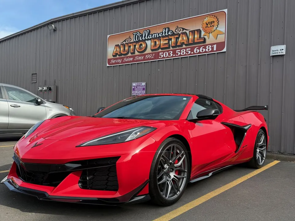 Red Corvette C8 with a tall rear wing parked in front of the shop's metal building