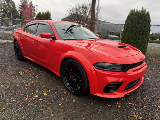 Red Dodge Charger with black wheels and a hood scoop parked on a gravel lot with fallen leaves