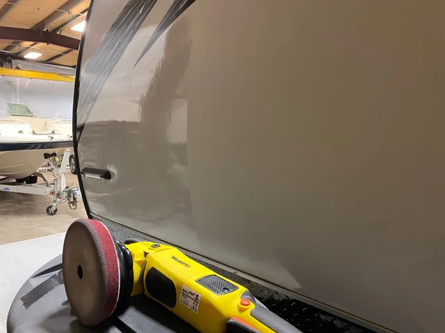 Yellow polisher with a red pad resting at the base of a gray RV wall, glossy on the left and dull on the right