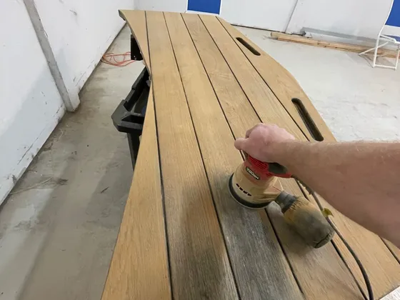 Hand running an orbital sander over a weathered teak slat panel, with the sanded part lighter than the gray part
