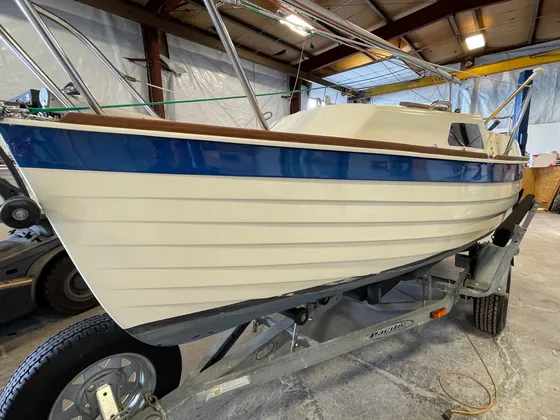 Small cream sailboat with a blue stripe and teak rail on a trailer inside the shop