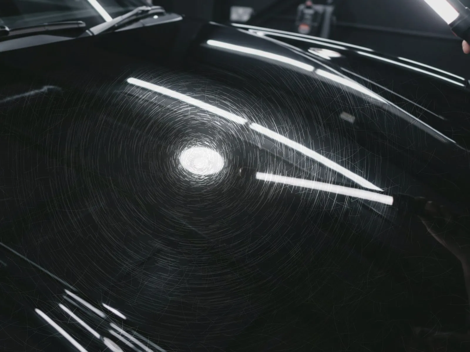 Black car hood covered in fine circular swirl marks that show around the reflection of an inspection light