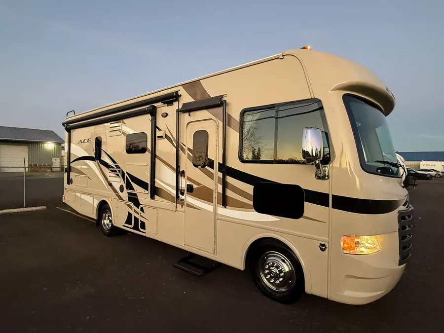 Tan ACE class A motorhome with black and brown graphics parked on a paved lot at dusk