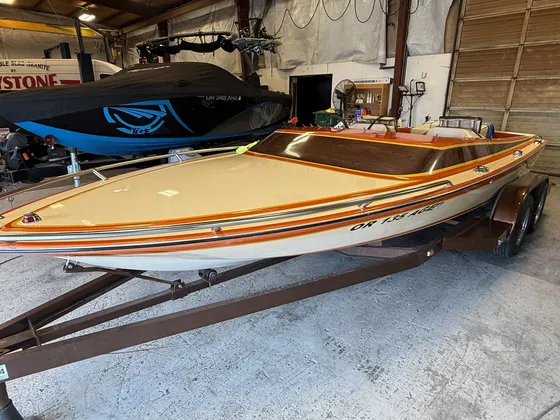 Older tan open-bow speedboat with orange stripes and a wood-look deck on its trailer inside the shop