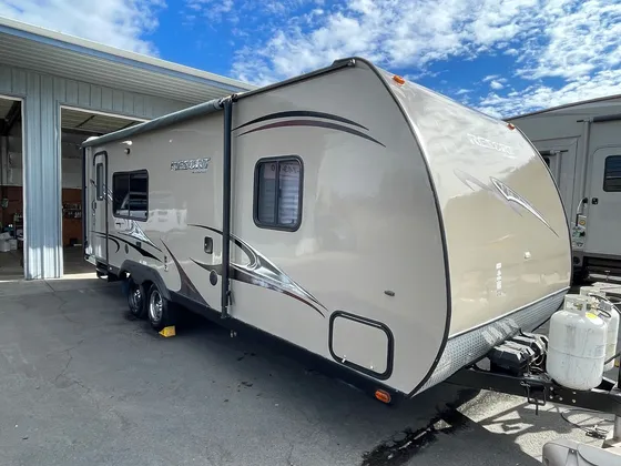 Tan Resort travel trailer with dark swoosh graphics and two propane tanks, parked by the shop door