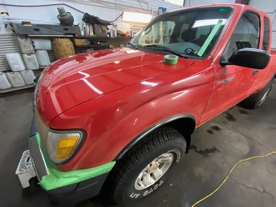 Red Toyota Tacoma extended-cab pickup with green masking tape on the bumper edge and windshield, tape roll on the hood