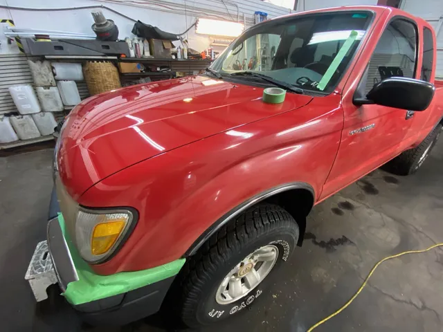 Red Toyota Tacoma extended-cab pickup with green masking tape on the bumper edge and windshield, tape roll on the hood