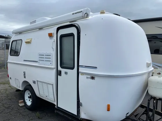 Small white molded fiberglass camper trailer with an awning and propane tank, parked in a lot