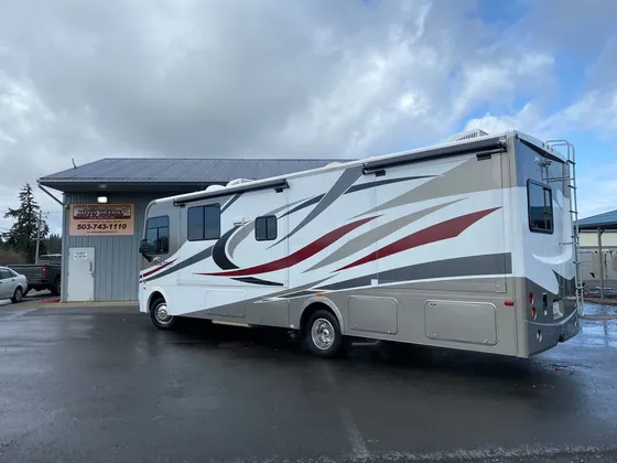 White and gray Class A motorhome with red and black swooshes parked outside the shop on wet pavement