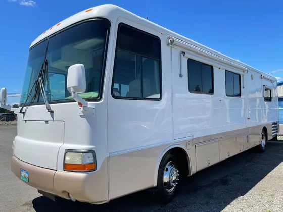 White and tan Class A motorhome seen from the front passenger corner on the lot under a blue sky