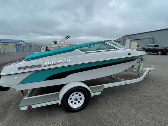 White runabout with teal stripe and teal bimini top on a white trailer outside under a cloudy sky